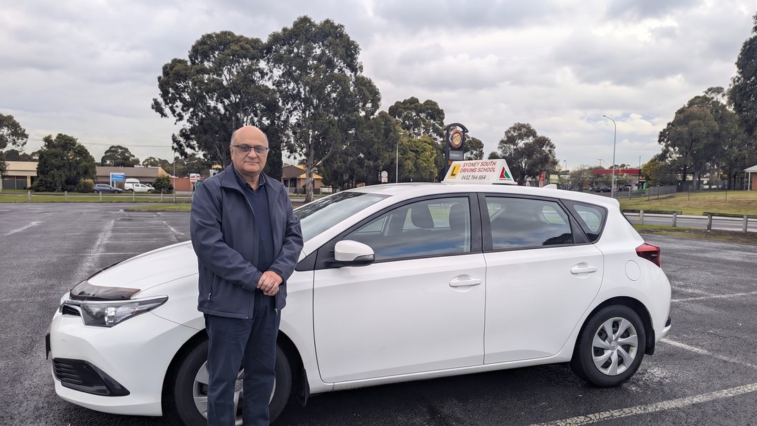 Ashraf George standing beside the white Sydney South Driving School dual-control automatic hatchback, with the roof sign showing the school name and phone number