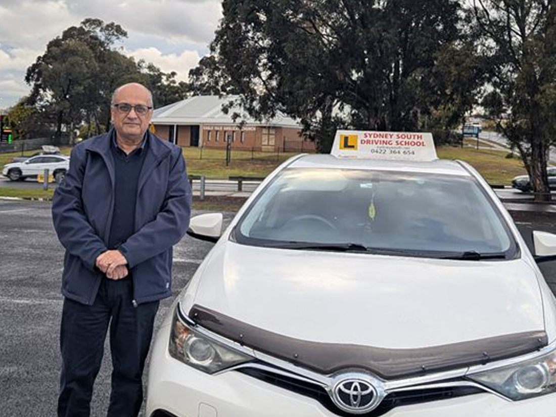 Ashraf, senior driving instructor at Sydney South Driving School, standing beside the school's white Toyota with its L-plate roof sign
