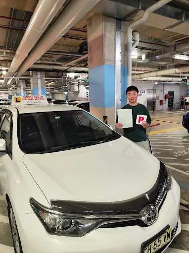 Student holding red P plates beside a white car in a car park after passing the driving test